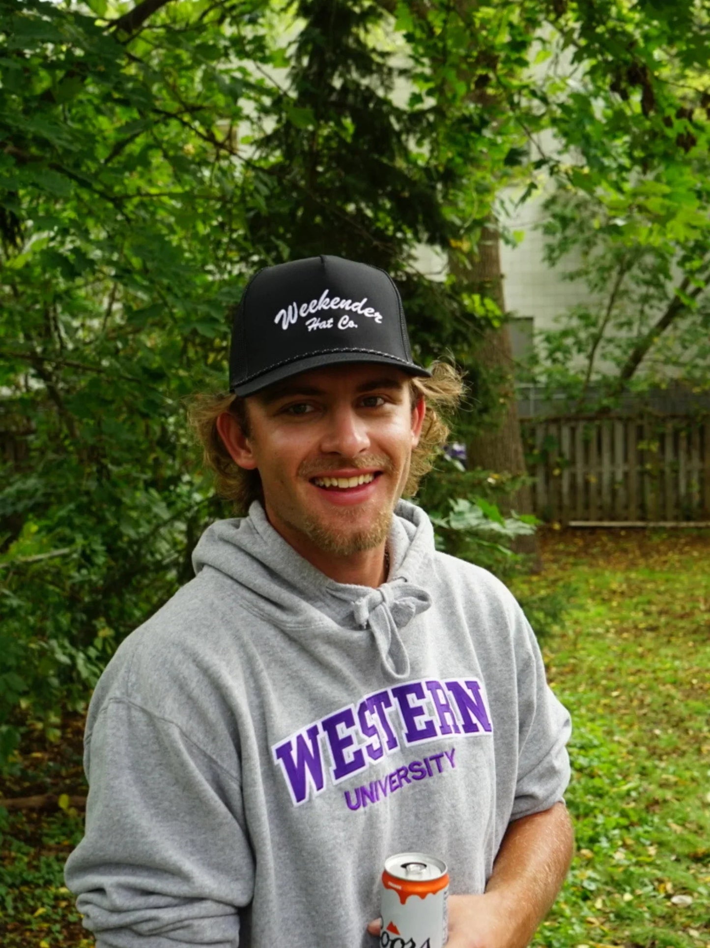 A photo of The Classic Weekender - Black Trucker Hat being worn in the backyard with a drink