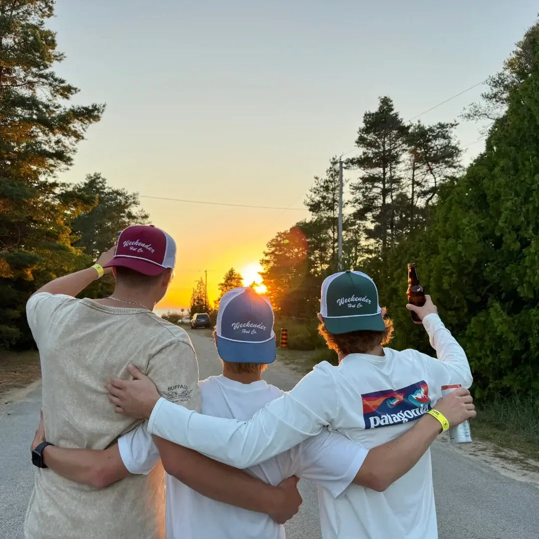 Red, blue, and green trucker hats from 'The Classics' collection watching sunset with drinks in hand - Weekender Hat Co.